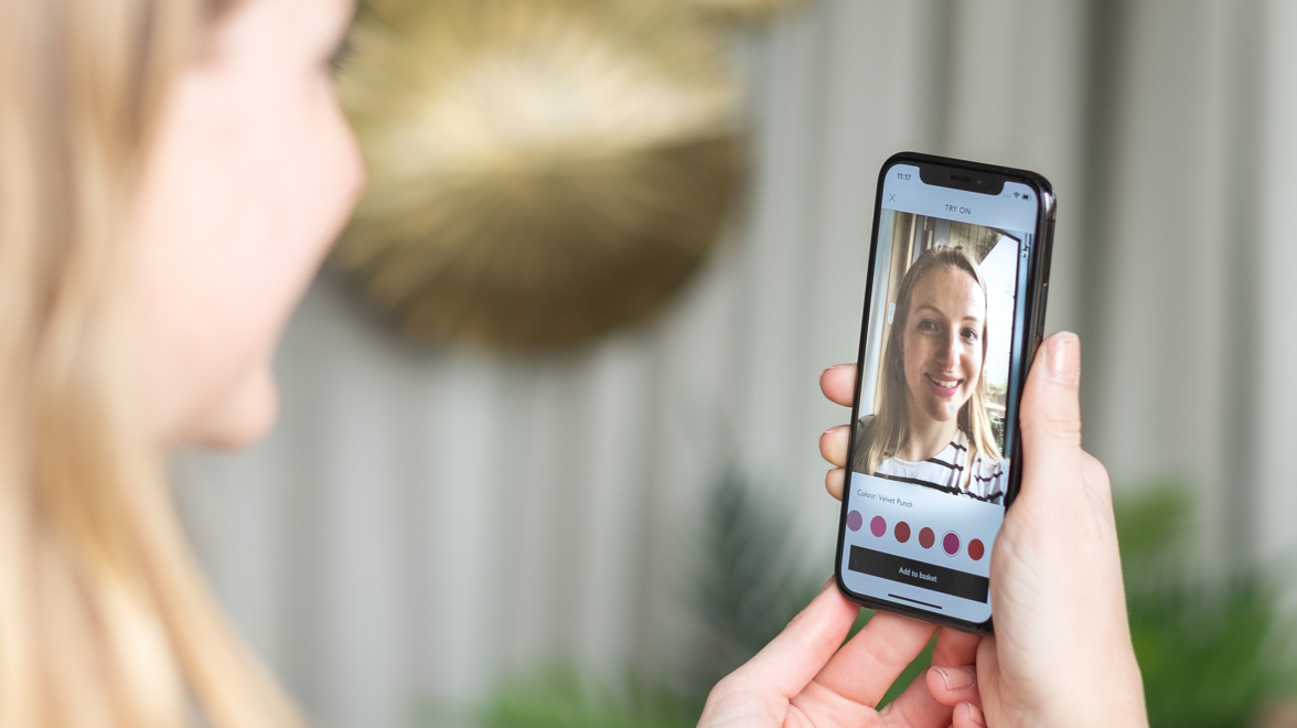 Someone holds a phone running the Lipstick try-on feature: the front camera shows their face with a shade applied, above a row of colour swatches and an Add to basket button.